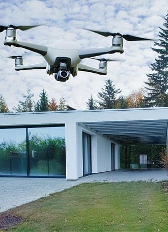 Drohne fliegt vor einem modernen weißen Haus mit großen Glasfenstern und umgebendem Garten.