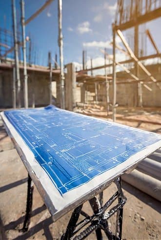 Bauplan auf einem Tisch vor einer Baustelle mit Gerüsten und teilweise errichteten Strukturen bei sonnigem Wetter.