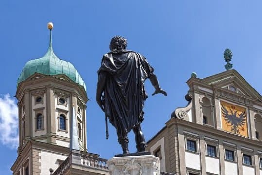 Denkmal von Kaiser Augustus vor dem Rathaus von Augsburg, flankiert von Türmen und unter einem klaren blauen Himmel.