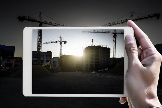 Hand hält Tablet mit Baustellenfoto, im Hintergrund Kran und Gebäude im Bau bei Sonnenuntergang.