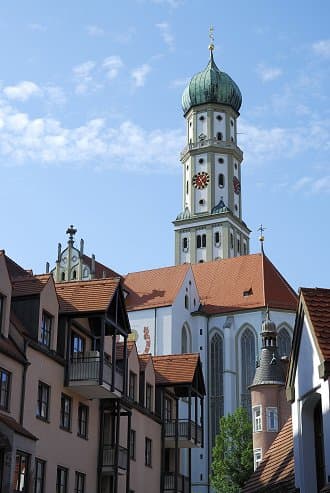 Blick auf den Turm der St. Ulrich und Afra Kirche in Augsburg, umgeben von traditionellen Häusern mit roten Dächern unter einem blauen Himmel.